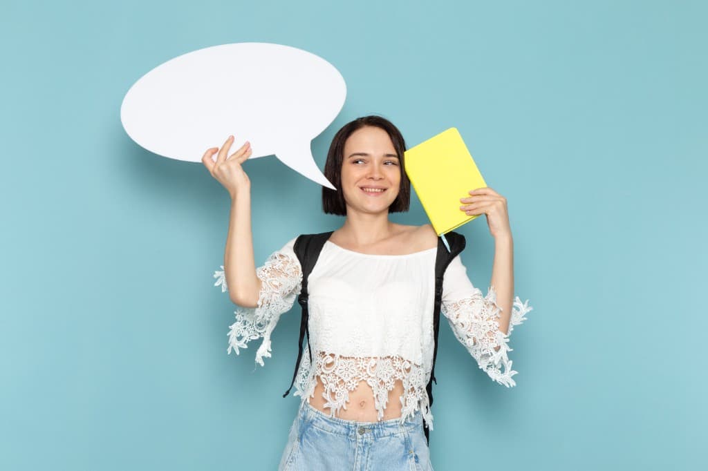 Smiling student holding a speech bubble sign and a yellow book on a soft blue background, representing campus voice on Kampus Connect